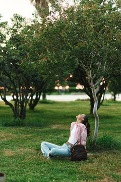 Student woman relaxes in a park, sitting against a tree with a backpack, wearing a striped shirt and jeans, enjoying a peaceful study break in nature during late afternoon
