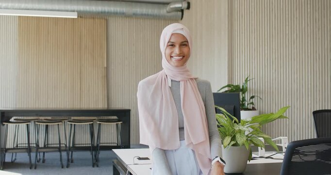 Adult woman wearing pink scarf engaging camera, leaning on office desk near plant and screen