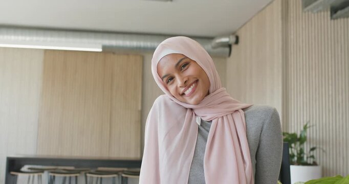 Adult woman with light pink headscarf smiling tilting left posing for camera in wood-paneled office