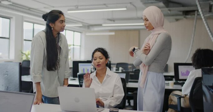 Diverse female staff joining seated woman at desk demoing on laptop while peer checking smartwatch