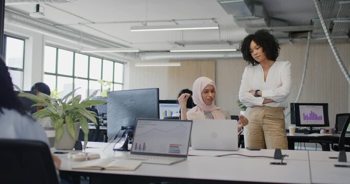 Diverse female coworkers approaching desk in office and discussing laptop charts to clarify data