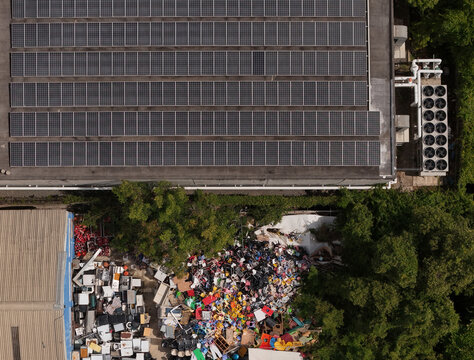 Aerial view of solar panels on a commercial building roof next to a cluttered recycling yard with waste materials and industrial equipment in Santo Domingo, Distrito Nacional, Dominican Republic.