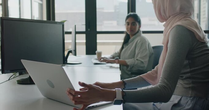 Diverse female coworkers working at desk, colleague pointing, hijab woman demonstrating on laptop