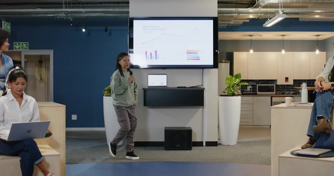 Nonbinary Indian speaker in sage top holding mic walking center in office before slide giving talk