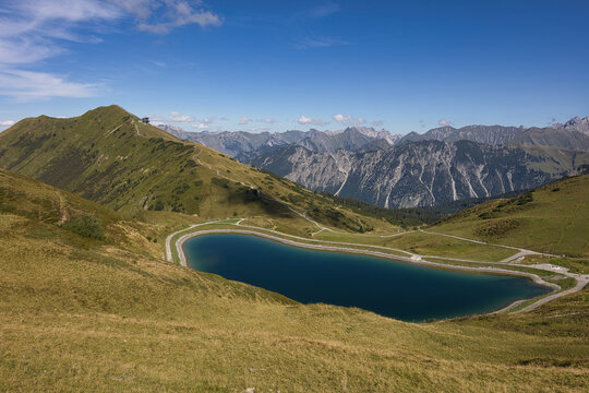 Ein idyllischer Bergsee liegt eingebettet in sanfte, goldgr&uuml;ne Almwiesen, umgeben von majest&auml;tischen Alpengipfeln unter einem strahlend blauen Himmel