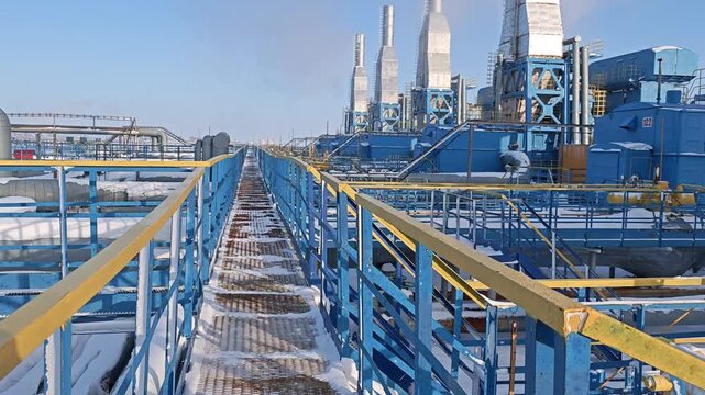Winter maintenance access continues along frosty metal walkway beside blue plant machinery. Snow lines grating and railings while exhaust stacks rise above utility complex, with steel stairs nearby.