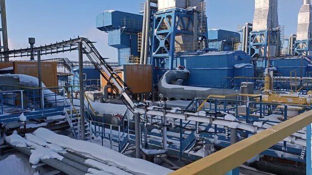 Frozen industrial plant shows blue towers, ducts, and catwalks covered by snow. Winter facility feels still and cold, with exposed machinery and raised walkways adding clear safety risk.