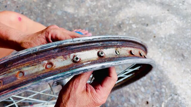 Close-up of a mechanic inspecting a heavily rusted motorcycle wheel rim with spokes outdoors. Concept of vintage bike restoration, wheel repair, worn metal parts, and maintenance work.