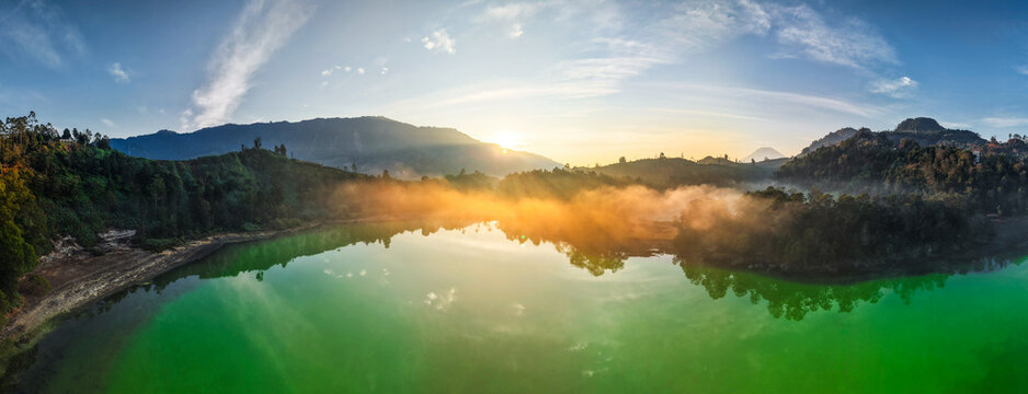 Aerial view of Telaga Warna Dieng lake with vibrant green water, surrounding hills, and morning mist at sunrise in Indonesia.
