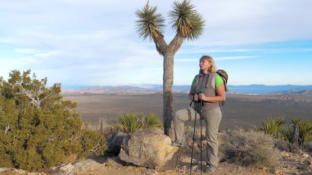 Mature woman with backpack and trekking poles actively hiking along narrow dirt path in rugged, arid hills landscape enjoying an adventurous journey through dry sunny wilderness. Hiking activities