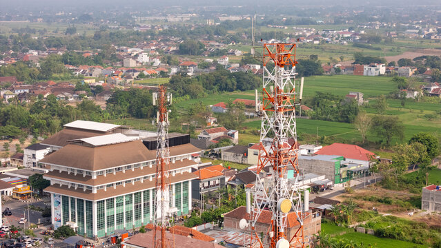 Aerial view of telecommunication towers rising above urban buildings and lush green fields in Klaten, Central Java, Indonesia.