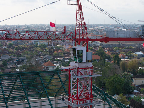 Aerial view of the Wangaya Regional General Hospital Emergency Room construction site with a red tower crane and Indonesian flag over urban rooftops in Denpasar Utara, Bali, Indonesia.