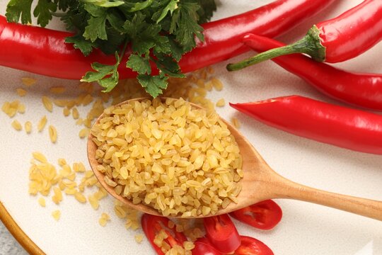 Raw bulgur in spoon, chili peppers and parsley on plate, top view