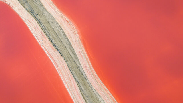 Aerial view of a diagonal access road crossing vivid red and pink salt evaporation ponds in Camargue, creating a geometric pattern in Aigues-Mortes, Occitanie, France.
