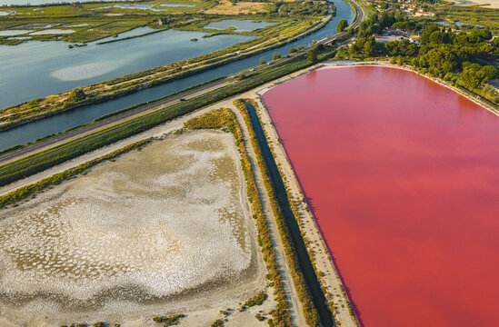 Aerial view of vivid red salt evaporation pond beside dry saline ground and water channel, contrasting landscape shaped by industrial sea salt production in Aigues-Mortes, Occitanie, France.