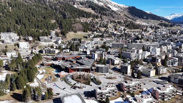 davos congress centre roundabout anchors dense alpine townscape switzerland snowcapped peaks tower over forested hillside slopes graubunden valley aerial drone footage wide establishing view clear sky