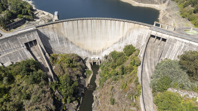 Aerial view of the massive concrete structure of the Belesar Dam surrounded by lush green forests and the deep waters of the Mino River in Lugo, Lugo, Spain.
