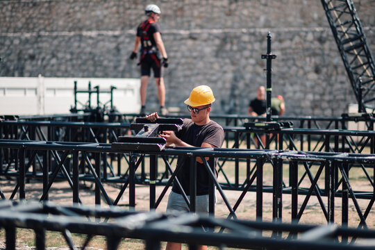 Worker in safety helmet and sunglasses carefully connecting parts of a temporary stage frame for an upcoming event