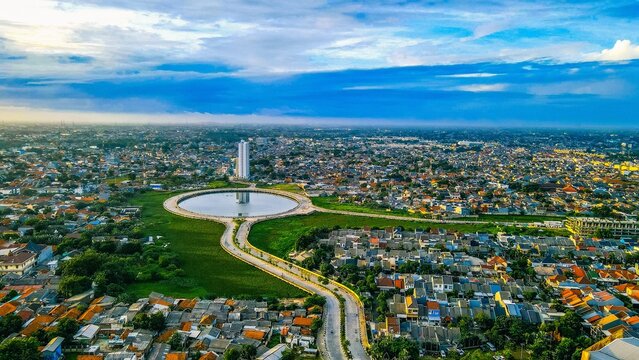 Aerial view of the circular lake and surrounding urban landscape with dense rooftops and a tall building in Bekasi Barat, West Java, Indonesia.