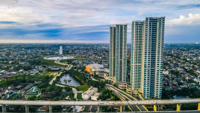 Aerial view of modern skyscrapers rising above the urban landscape of Bekasi Barat with a highway overpass, a reflective pond, and residential rooftops, Bekasi Barat, West Java, Indonesia.