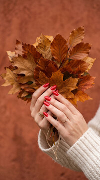 Woman's hands hold a bouquet of dried autumn leaves, showcasing red polished nails and rings, symbolizing fall and nature
