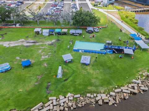Wilmington, United States - 12 July 2025: Aerial view of an outdoor event with a temporary swimming pool and vendor tents on a grassy field near the Battleship North Carolina.