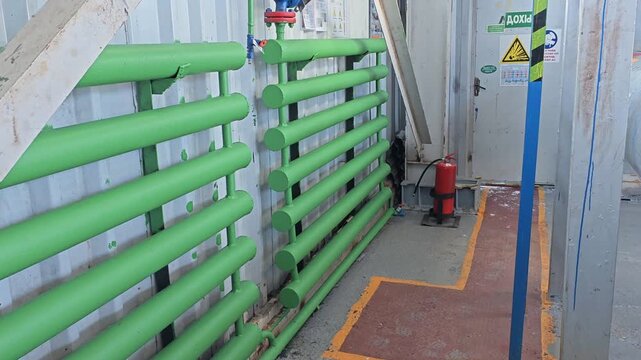 Large insulated pipe assembly dominates industrial maintenance space. Green pipe rack and blue flanges sit beside warning signs, creating functional utility setting with clean lines and workmanlike