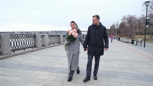 Romantic couple walking along riverside promenade while woman carries bouquet of pink roses