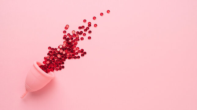 Pink menstrual cup with red sequins spilling out on a soft pastel pink background flat lay