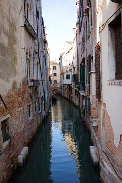 A distant gondola glides down a narrow Venetian canal beneath brick buildings and a small stone bridge.