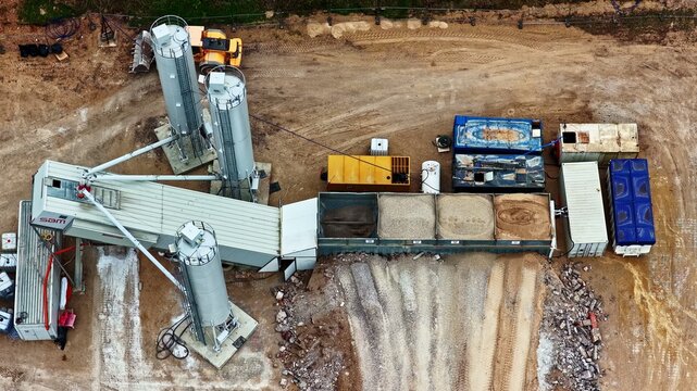 Valmiera, Latvia - 28 May 2026: Aerial view of a concrete batching plant with industrial silos, aggregate storage bins, and construction machinery on a dirt site.