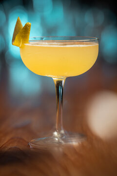 Elegant cocktail, the Bees Knees in coupe glass. Lemon twist adorns the rim. Warm wood table contrasts cool background. Soft bokeh lights enhance drink's allure. Perfect for bar, lounge