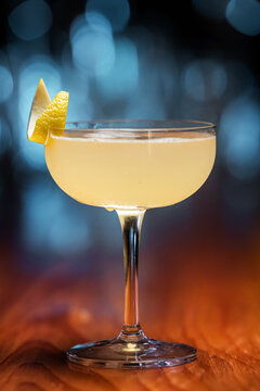 Elegant cocktail, the Bees Knees in coupe glass. Lemon twist adorns the rim. Warm wood table contrasts cool background. Soft bokeh lights enhance drink's allure. Perfect for bar, lounge