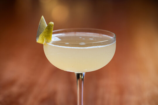 Elegant cocktail, the Bees Knees in coupe glass. Lemon twist adorns the rim. Warm wood table. Perfect for bar, lounge