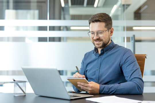Professional businessman working on laptop and taking notes in modern office, remote work and corporate productivity, Focused male employee using computer and writing in notebook for business