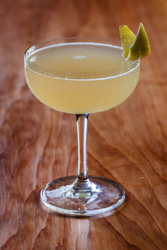 Elegant cocktail, the Bees Knees in coupe glass. Lemon twist adorns the rim. Warm wood table. Perfect for bar, lounge
