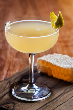 Elegant cocktail, the Bees Knees in coupe glass. Lemon twist adorns the rim. Warm wood table and honey comb garnish.