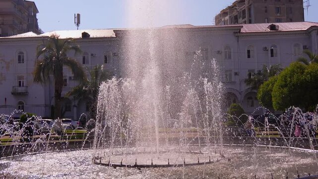 Water jets spray from a large circular fountain in a public park on a sunny day, while an old classical building and palm trees create a tranquil cityscape in Kobuleti in Georgia