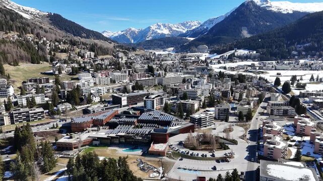 snowcapped peaks tower over davos valley switzerland congress centre timber roof anchors foreground dense rooftops chalets climb forested hillside aerial drone orbit wide establishing clear sky