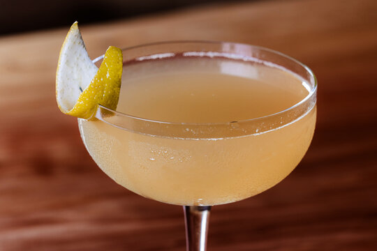 Elegant cocktail, the Bees Knees in coupe glass. Lemon twist adorns the rim. Warm wood table. Perfect for bar, lounge