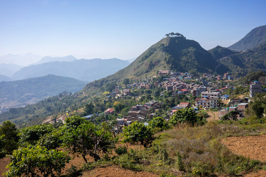 Elevated daytime view of a hillside village near Bandipur, Nepal, with houses scattered across a forested hill under a clear blue sky.