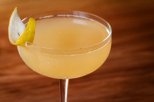 Elegant cocktail, the Bees Knees in coupe glass. Lemon twist adorns the rim. Warm wood table. Perfect for bar, lounge