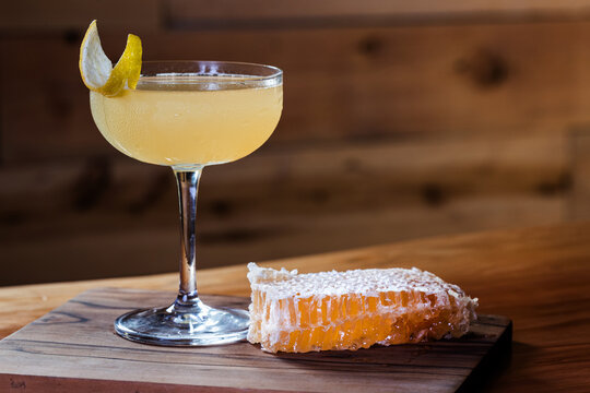 Elegant cocktail, the Bees Knees in coupe glass. Lemon twist adorns the rim. Warm wood table and honey comb garnish.