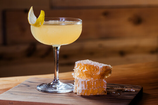 Elegant cocktail, the Bees Knees in coupe glass. Lemon twist adorns the rim. Warm wood table and honey comb garnish.