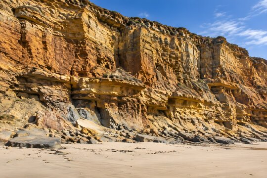 Warm sandstone cliff rising above a sandy beach with banded ledges and fallen slabs