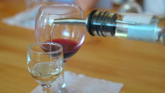 Close-up view of a bartender pouring white wine from a bottle with a pourer spout into a small tasting glass, with another glass of red wine sitting on the wooden table in the background