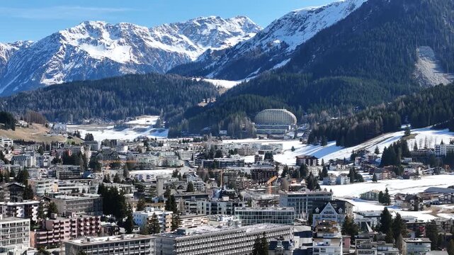 forested ridge frames davos valley floor switzerland domed roof building nestled among pines rooftops spread below snowcapped mountain range towers behind aerial telephoto static hover clear sky