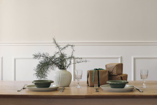 Minimalist Christmas Table Setting with Green Ceramic bowls, Dinnerware and drinking glass. Wrapped Gift boxes, pine tree branches in vase. Elegant winter home decor, Scandinavian interior.