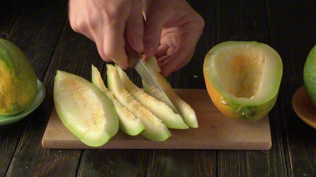 Hands slicing green melon into pieces on wooden cutting board, with whole melons and a bowl in the background, showcasing the preparation process in a kitchen setting