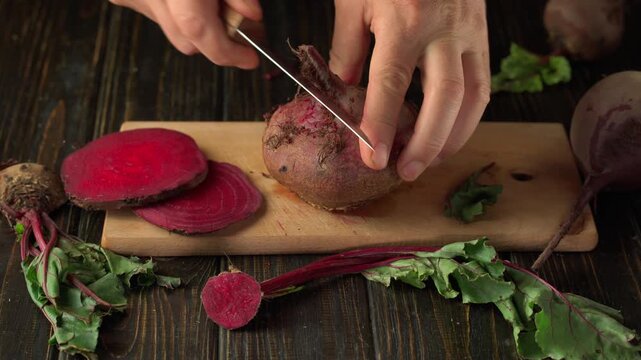 Hands skillfully peel a beetroot on a wooden board, surrounded by bright red beetroot slices and green leaves, highlighting the process of preparing fresh vegetables in the kitchen